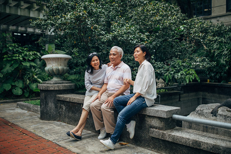 Two ladies sitting with their elderly father on a bench